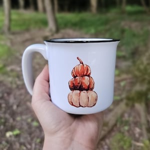 Könnte beinhalten: Weißer Keramikbecher mit schwarzem Rand und Henkel. Der Becher zeigt eine Aquarellillustration von drei gestapelten Kürbissen in Orange- und Beigetönen. Der Becher wird in einer Hand vor einem verschwommenen grünen Hintergrund gehalten.