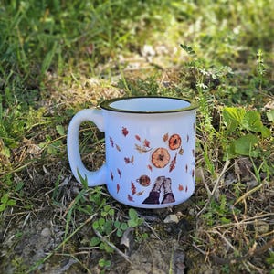 Könnte beinhalten: Weißer Emaille-Becher mit schwarzem Rand und Henkel, verziert mit Herbstblättern, Kürbissen und einer gemütlichen Illustration von zwei Personen. Der Becher steht auf einem Bett aus grünem Gras und kleinen Pflanzen.