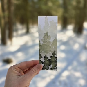 Può includere: Un segnalibro ad acquerello con una scena di foresta nei toni del verde e del grigio. L'opera raffigura alberi sempreverdi stilizzati, con uno sfondo sfocato che suggerisce un paesaggio innevato. Il segnalibro è tenuto su uno sfondo di alberi e neve.