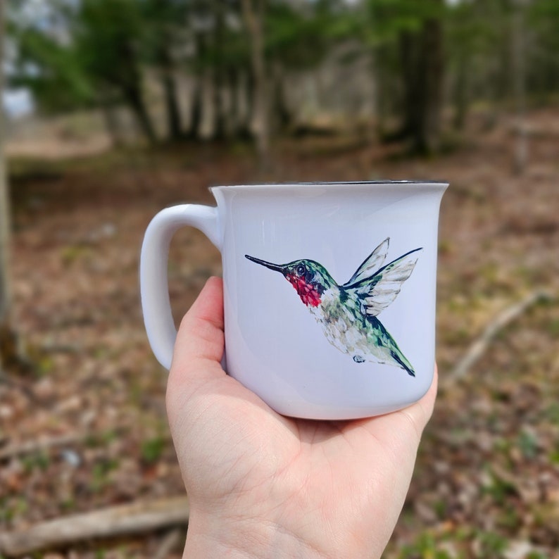 May include: White ceramic mug with a black rim and handle. The mug features a watercolor illustration of a hummingbird with green and red accents, flying to the right.