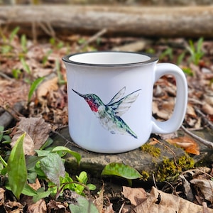 May include: White ceramic mug with a black rim and handle. The mug features a watercolor illustration of a hummingbird in flight with green, red, and black colors.