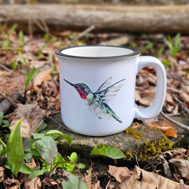 May include: White ceramic mug with a black rim and handle. The mug features a watercolor illustration of a hummingbird in flight with green, red, and black colors.