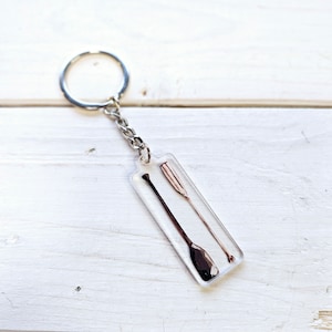 Peut inclure: Un porte-clés transparent avec une chaîne argentée et un charme rectangulaire contenant deux pagaies en bois à l'intérieur.