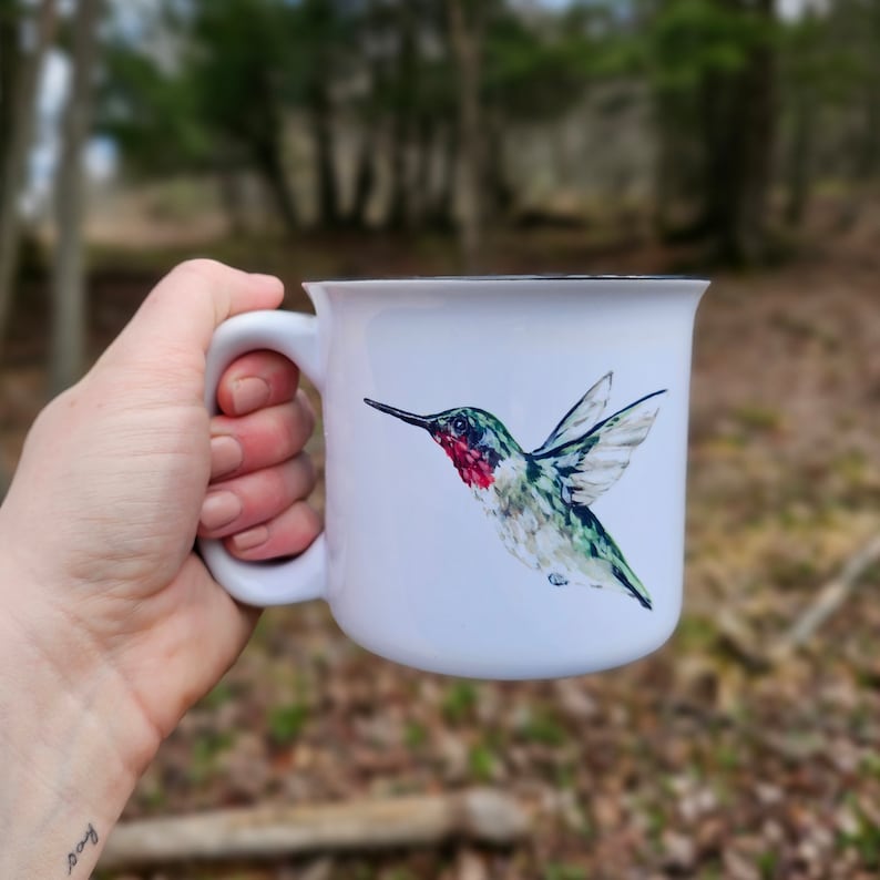 May include: White ceramic mug with a hummingbird illustration. The hummingbird is green, red, and black with a long beak and outstretched wings.