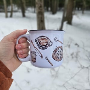 May include: White enamel mug with a black rim featuring a watercolor illustration of bread baking tools and loaves of bread. The mug is being held by a hand in a brown jacket.