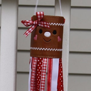 May include: A whimsical gingerbread man wind chime. The brown cylindrical body features a smiling face, heart cheeks, and a red and white gingham bow. Red, white, and gingham ribbons cascade down, creating a festive, decorative piece.