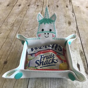 May include: White felt snack tray with a unicorn applique. The unicorn has a green mane and pink hearts for cheeks. The tray has turquoise buttons on the sides. The tray is holding a small package of fruit snacks.