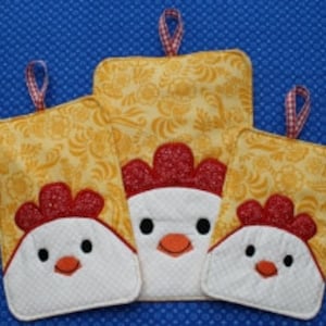 Pode incluir: Três pegadores de panela amarelos e brancos com um design de frango. Os pegadores de panela são de tamanhos diferentes e têm acentos vermelhos.