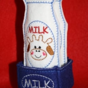 May include: A felt toy milk carton with two felt milk bottles inside. The milk bottles are white with blue stitching and the word "MILK" embroidered on the front. The carton is blue with the word "MILK" embroidered on the front. A cartoon cow face is embroidered on the front of the milk bottles.