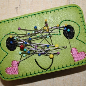 Könnte beinhalten: Ein grünes Leder-Nadelkissen in Form eines Frosches mit rosa Herzen und schwarzen Augen. Das Nadelkissen ist mit bunten Stecknadeln gefüllt.