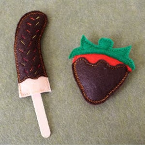 May include: Two felt food toys, a chocolate-covered popsicle and a chocolate-covered strawberry. The popsicle is pink and white with a brown stick. The strawberry is red with a green stem and leaves.