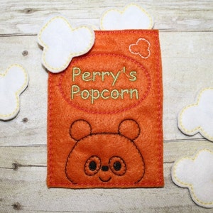 Puede incluir: Bolsa de fieltro naranja con una cara de panda bordada en negro y el texto "Perry's Popcorn". Cinco piezas de palomitas de maíz de fieltro blanco están dispersas alrededor de la bolsa.
