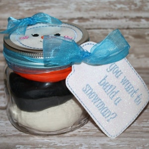 May include: A glass jar with a white, black, and orange play dough inside. The jar is decorated with a blue ribbon and a snowman-shaped lid. A white tag with the text "You want to build a snowman?" is attached to the jar.