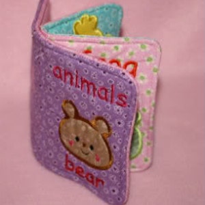 May include: A purple and pink fabric baby book with a bear on the front page. The book has the word "animals" and "bear" embroidered on the front page.