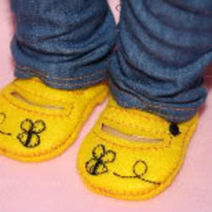 Puede incluir: Primer plano de zapatos de fieltro amarillo con diseño de abeja y vaqueros azules. Los zapatos tienen una correa en la parte superior y un diseño de abeja y remolino cosido. Los vaqueros están doblados en los tobillos.