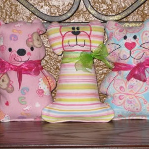 May include: Three plush toys, a pink bear, a striped dog, and a blue cat, all with embroidered faces and pink and green ribbons.