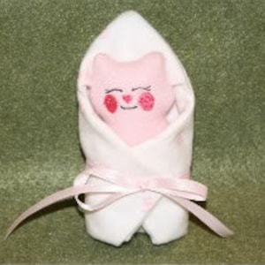 May include: A pink and white baby doll swaddled in a white blanket with a pink ribbon. The doll has a pink face with red cheeks and embroidered eyes.