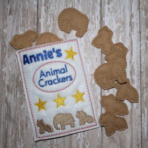 May include: A white felt bag with the text "Annie's Animal Crackers" and three yellow stars. The bag is filled with felt animal crackers, including an elephant, a camel, and a bear.