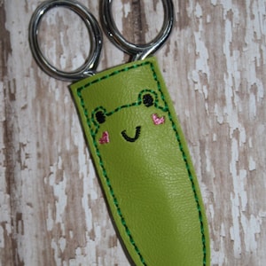 Può includere: Un astuccio per forbici in pelle verde con un simpatico viso di rana ricamato. La rana ha occhi neri, un sorriso nero e guance rosa.
