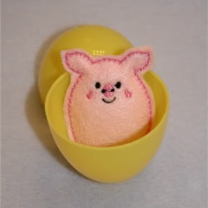 May include: A light pink felt pig plushie with embroidered details, including a smiling face and heart-shaped cheeks, sits inside a bright yellow plastic egg. The egg is open, revealing the pig. The background is a light blue.