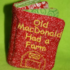 May include: Red fabric book with embroidered text "Old MacDonald Had a Farm" and a red barn with a yellow door.