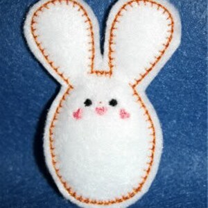 Könnte beinhalten: Ein weißer Filzhase mit braunen Nähten an den Rändern und einem einfachen gestickten Gesicht mit schwarzen Augen und rosa Wangen.