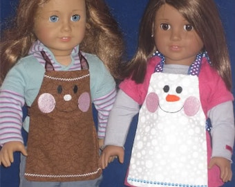 May include: Two dolls wearing aprons. One doll has a brown apron with a gingerbread man design, featuring pink cheeks and a smiling face. The other doll wears a white apron with a snowman design, also with pink cheeks and a smiling face.
