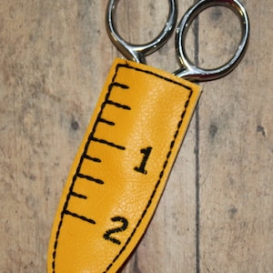 Può includere: Un astuccio per forbici in pelle gialla con un design a righello cucito in nero. L'astuccio ha i numeri "1" e "2" ricamati. Un paio di forbici argentate sporgono dalla parte superiore dell'astuccio.
