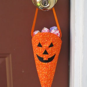 May include: An orange fabric Halloween treat bag shaped like a jack-o'-lantern, hanging from a doorknob. The bag has a black face and is filled with wrapped candies. The bag is made of orange fabric with a pattern of small orange and yellow shapes.