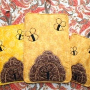Puede incluir: Conjunto de tres agarraderas de olla amarillas y marrones con un diseño de colmena y abejas. Las agarraderas de olla están hechas de tela y tienen un lazo para colgar.