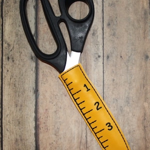May include: Black scissors with a yellow fabric ruler cover. The ruler markings are in inches and go up to 3 inches.