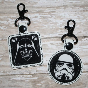 May include: Two black and white embroidered keychains featuring Star Wars characters. The first keychain is square and features Darth Vader's face. The second keychain is round and features a Stormtrooper's face.