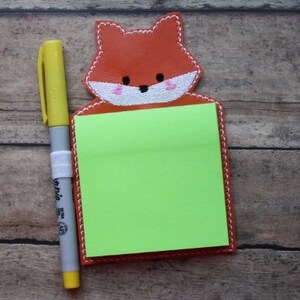 Op de afbeelding: Een bruine, vosvormige sticky note-houder met een groene sticky note erin. Een gele en grijze marker ligt naast de houder.
