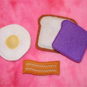 Może przedstawiać: Zestaw filcowej żywności do zabawy z białym jajkiem, dwoma kromkami białego chleba, kromką fioletowego dżemu i kromką brązowego bekonu.