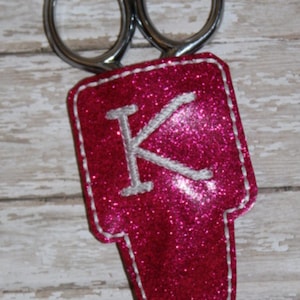 May include: A pink glitter scissor holder with white stitching and the letter "K" embroidered in white.