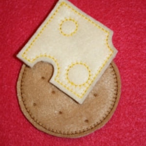 May include: A felt craft of a round brown cracker with a square piece of white felt cheese on top. The cheese has two holes and a bite taken out of it.