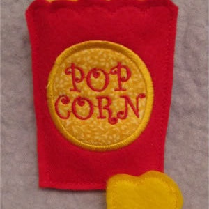 Puede incluir: Una bolsa de palomitas de maíz de fieltro rojo con una pieza de palomitas de maíz de fieltro amarillo y un parche bordado de fieltro amarillo que dice "POP CORN".