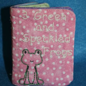May include: A pink fabric book with white polka dots and the text "3 Green and Speckled Frogs" embroidered on the front cover. A green frog with white eyes is also embroidered on the cover.