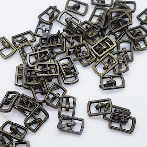 Puede incluir: Una gran cantidad de hebillas deslizantes de metal dorado antiguo. Las hebillas son todas del mismo tamaño y forma, y están esparcidas al azar sobre una superficie blanca.