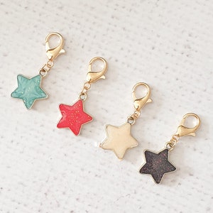May include: Four star-shaped charms in teal, red, cream, and black with gold-coloured clasps. Each charm has a gold-coloured border. The charms are displayed on a white, textured surface.
