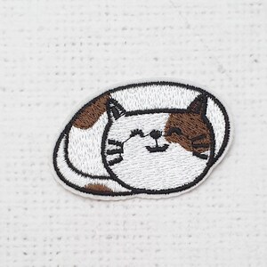 Puede incluir: Parche bordado con un gato durmiendo. El gato es blanco con manchas marrones y un contorno negro. El parche tiene forma ovalada y muestra una expresión sonriente. El fondo es una tela blanca texturizada.