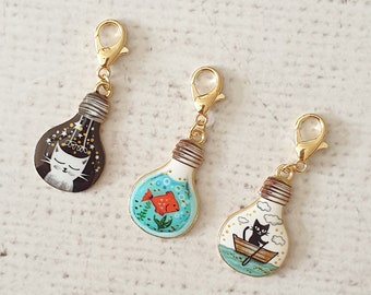 May include: Three gold lightbulb charms with different designs. The first charm features a white cat with stars above its head. The second charm features a red fish swimming in a blue and green ocean. The third charm features a black cat in a boat with clouds above it.