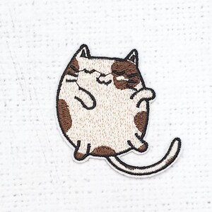 Puede incluir: Parche bordado con un gato de dibujos animados en una pose juguetona. El gato tiene un cuerpo blanco con manchas y marcas marrones. El parche está delineado en negro y tiene un fondo blanco.