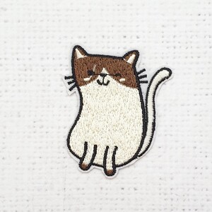 Puede incluir: Parche bordado con un gato sonriente de pelaje marrón y blanco. El gato tiene un contorno y bigotes negros. El parche está diseñado para planchar o coser sobre tela, ideal para personalizar ropa o accesorios.