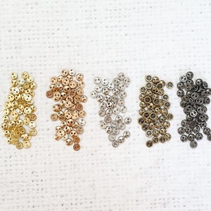 May include: Assortment of small, round buttons in four different metallic colours: gold, silver, brass, and black. The buttons are arranged in four rows on a white textured surface.