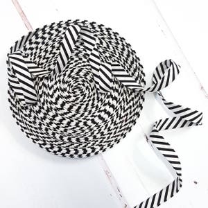 May include: A coiled roll of black and white patterned ribbon. The ribbon features a repeating geometric design and is partially unwound, revealing a striped edge. It rests on a white wooden surface.