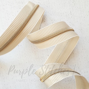 May include: A close-up of a beige zipper with a gold metal zipper pull. The zipper tape has a subtle striped pattern.