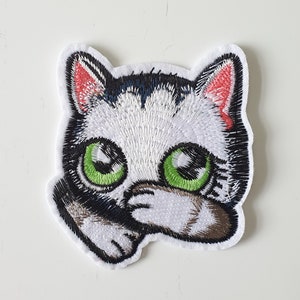 May include: Embroidered iron-on patch of a white and black cat with green eyes. The cat is looking to the right and has its paw covering its face.