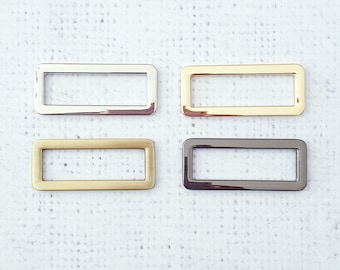 May include: Four rectangular metal buckles in silver, gold, brass, and gunmetal finishes. The buckles are arranged in a square pattern against a white textured background. These are likely used for bag making or other craft projects.