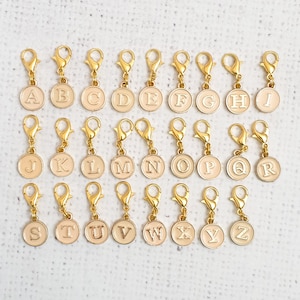 May include: Set of 26 gold-toned alphabet letter charms with a cream-colored background. Each charm features a different letter of the alphabet.
